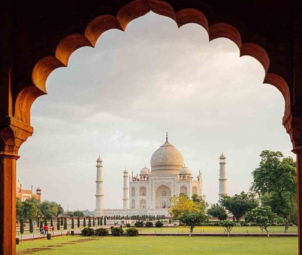 The Taj Mahal framed by an ornate arch, set against a soft sky. Lush green gardens in the foreground add to the serene scene.
