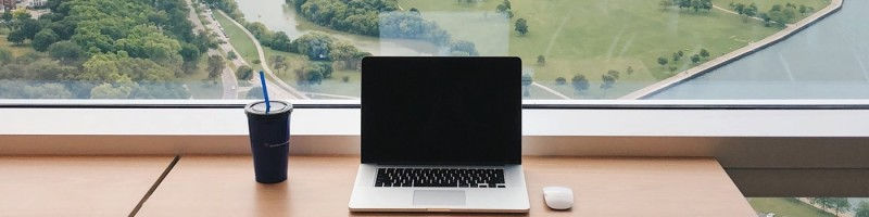 Laptop and cup on desk by large window, overlooking scenic park and river. Bright, serene setting with green trees and clear sky.