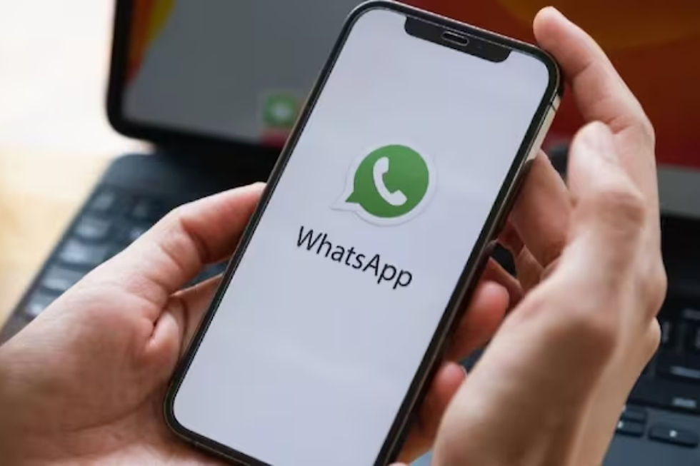 Hands holding a smartphone showing the WhatsApp logo and text. A laptop is in the blurry background, suggesting a tech setting.