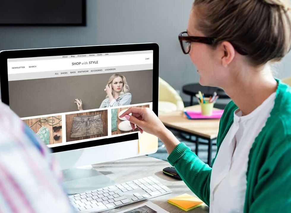 Person in glasses and green sweater points at clothing items on a computer screen in a modern office. The website reads "SHOP with STYLE."