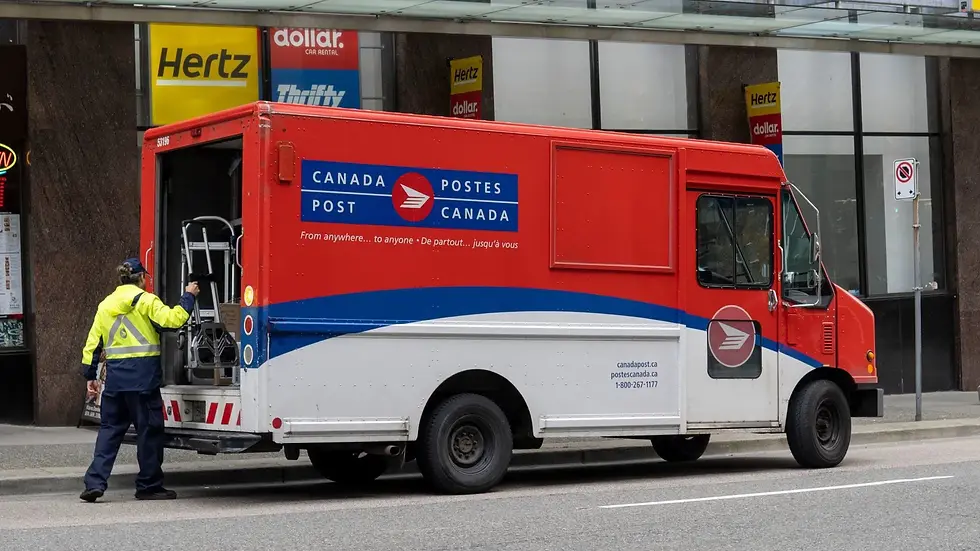 Canada Post truck with red and white colors parked on city street. Worker in yellow jacket loads packages. Hertz signs visible in background.