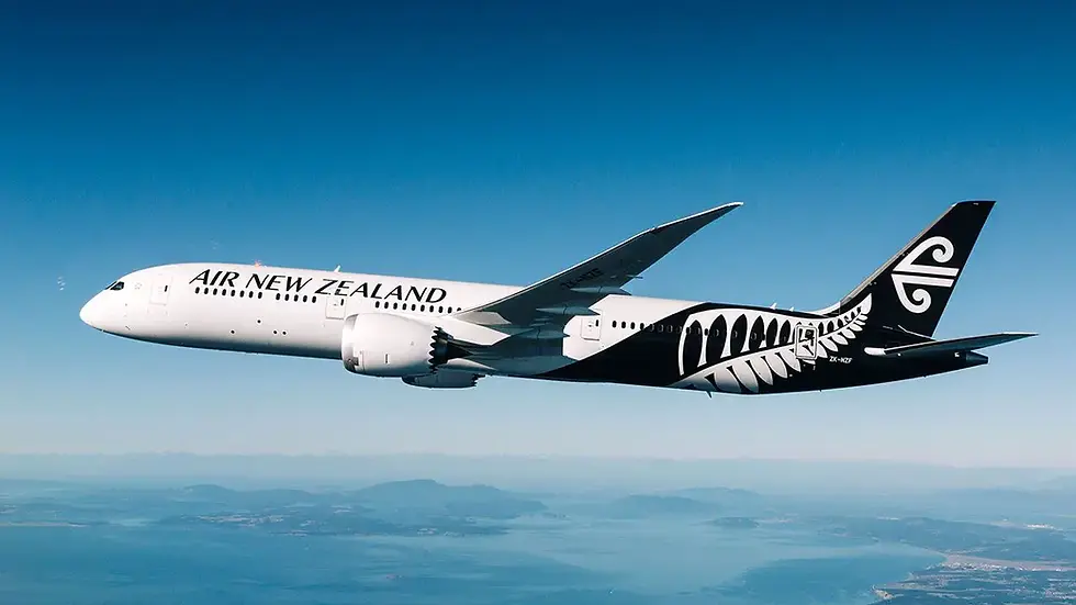 White Air New Zealand plane with black Maori design flies over ocean and distant land under clear blue sky.