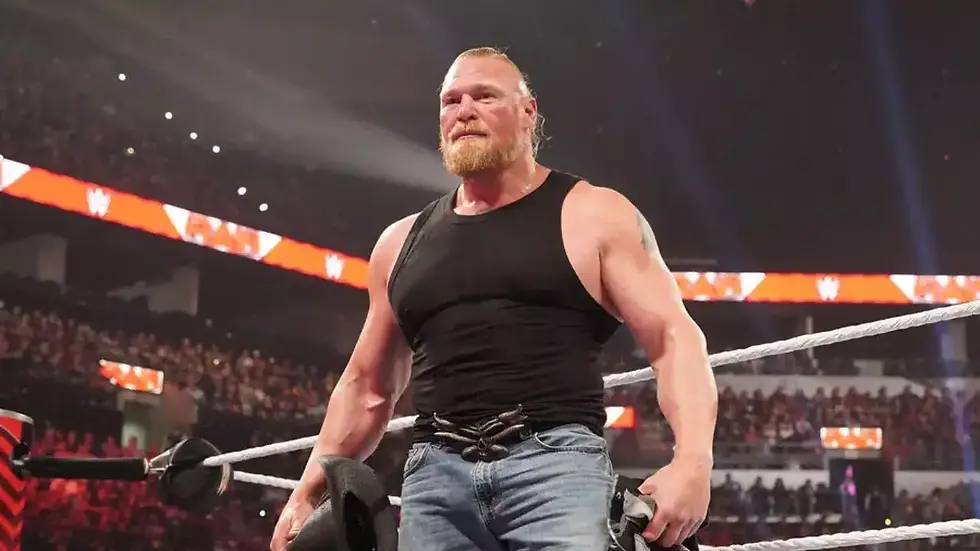 Wrestler in black tank top and jeans stands in a ring, holding a cowboy hat. Audience and red lights in the background create an intense mood.