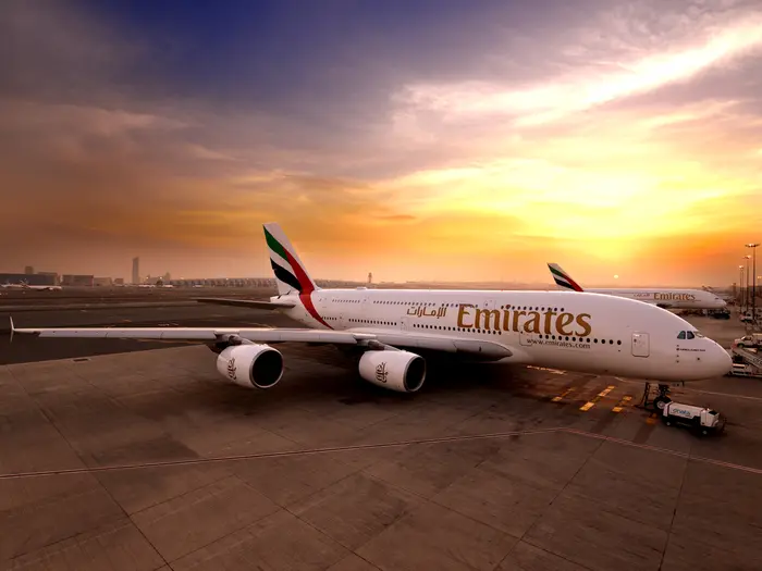 Emirates airplane parked at airport during a vibrant sunset, with an orange sky. Text "Emirates" visible on the plane. Calm, serene mood.