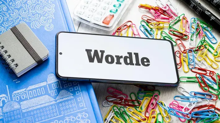 Smartphone displaying "Wordle" on screen surrounded by colorful paperclips, a notebook, a blue book, and a pillbox. Bright, cluttered setting.