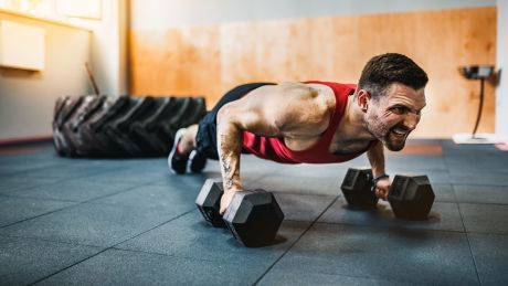 Man in a red tank top doing push-ups with dumbbells in a gym. Large tire in the background. Intense and focused expression.