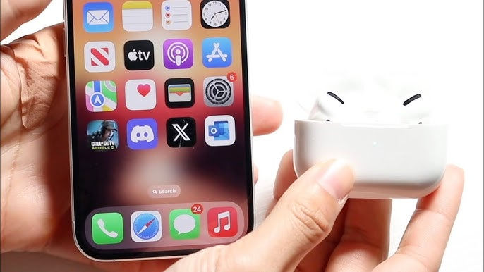 Hands holding a smartphone with colorful app icons and AirPods in a white case. Notification badges visible. Bright background.