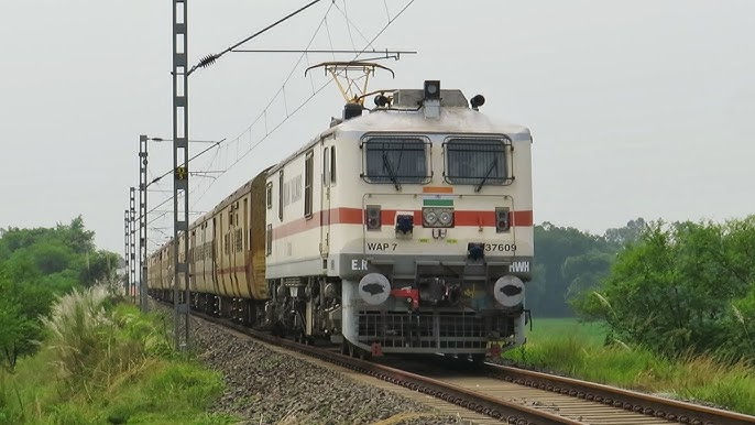 Electric train on tracks in a lush green landscape, powered by overhead cables. Train has a white and red color scheme, WAP 7 model.
