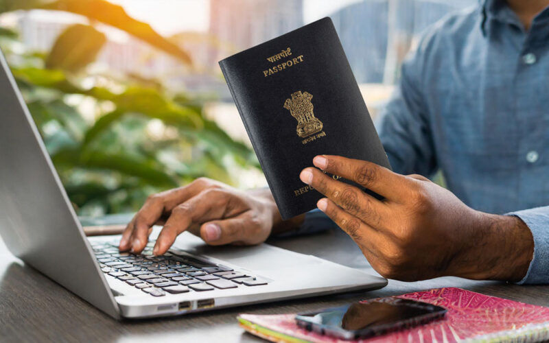 A person holds a passport while typing on a laptop in a bright room with plants. An ID and smartphone rest nearby on the wooden desk.