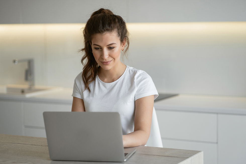 Woman in a white shirt typing on a laptop at a kitchen table. Background is modern and minimalistic. She looks focused and content.