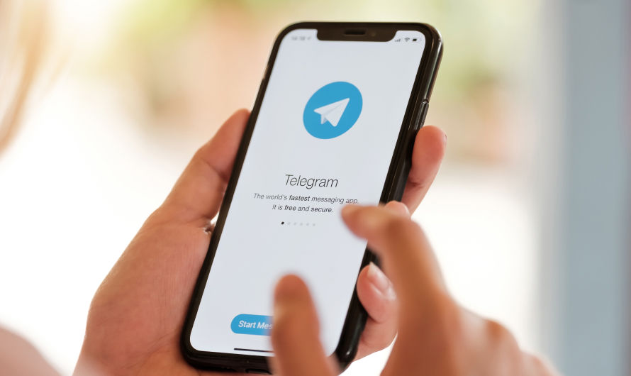 Close-up of a hand holding a phone showing the Telegram app welcome screen with text. The background is blurred and bright.