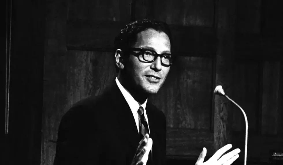 Man in suit and glasses speaks at a microphone, gesturing with hands. Dark wooden background; serious expression. Black and white image.