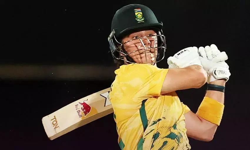 Cricketer in yellow jersey and helmet swings bat, dark background. Bat shows "TON" and "Red Bull" logos. Focused expression.