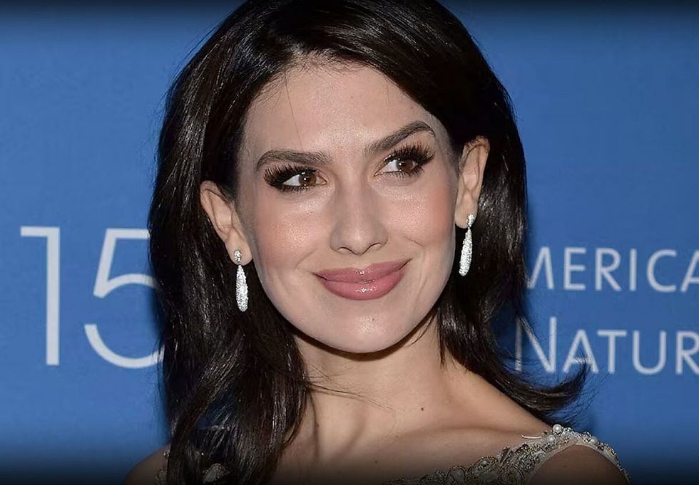 Woman smiling in front of a blue backdrop with "American" text. She wears sparkling earrings and an elegant dress, exuding a joyful mood.