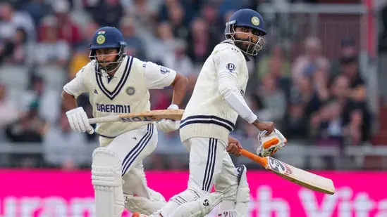 Two cricket players in white uniforms run between wickets on a grass field. One holds a bat. Crowd and pink advertisement board in background.