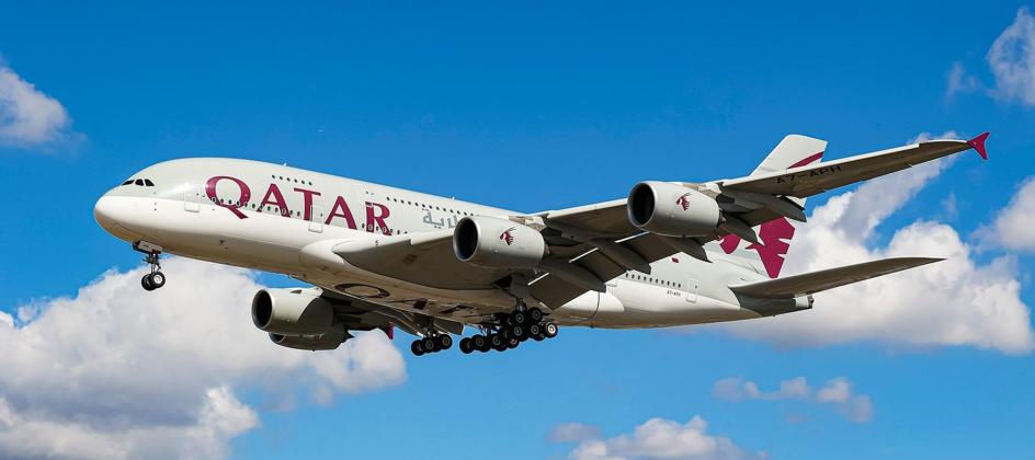 Qatar Airways plane flying against a bright blue sky with clouds. The aircraft is white with maroon text and accents, landing gear visible.
