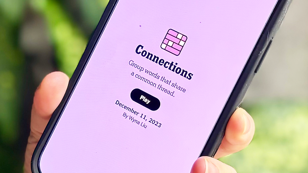 Close-up of a smartphone with the "Connections" game on a pink screen. Text includes "Group words," "Play," and date, held with a blurred green background.