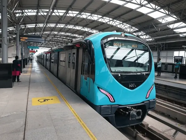 A bright blue metro train is stopped at a platform under a large arched roof. Passengers are nearby. The train display reads "0012 ALU".