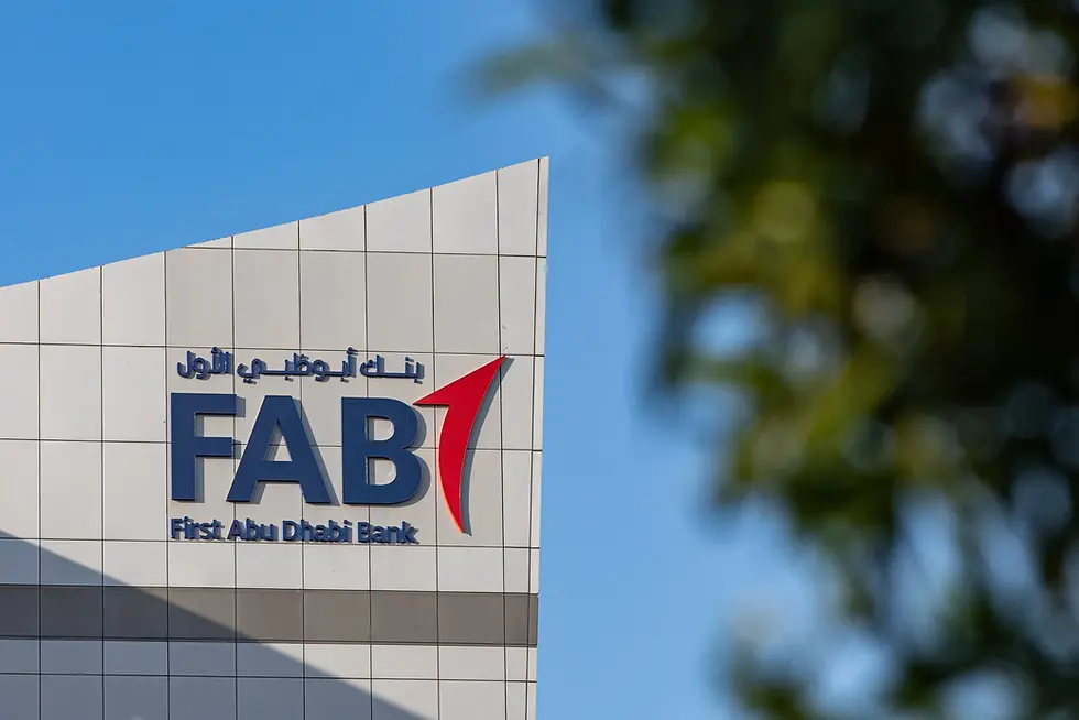Building facade with "FAB" and "First Abu Dhabi Bank" logo in blue and red. Clear blue sky and blurred greenery in the background.