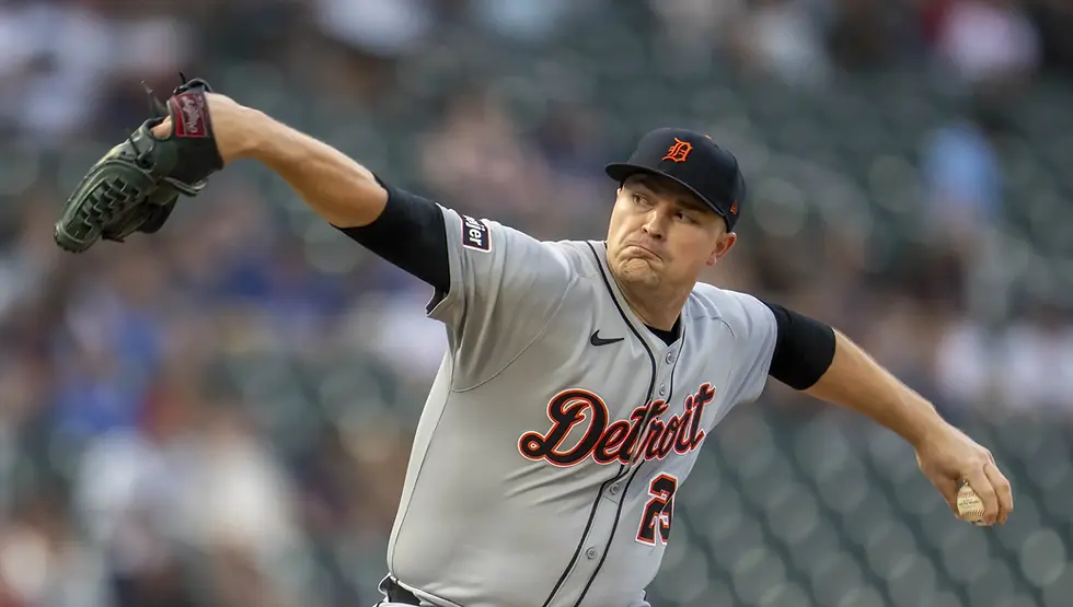 Baseball pitcher in a gray "Detroit" jersey throws a pitch with focus. He wears a black cap, in a stadium with blurred audience.