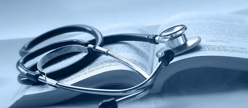 A stethoscope rests on an open book with visible text. The image has a blue tint, suggesting a medical or educational setting.