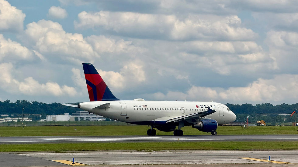 Airplane with Delta logo taxis on a runway under a cloudy sky. Grass and airport buildings are visible in the background.