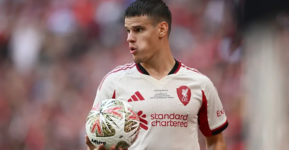 Soccer player in white and red LFC jersey holds a Mitre ball, mouth open, on a blurred stadium background, text reads "standard chartered".