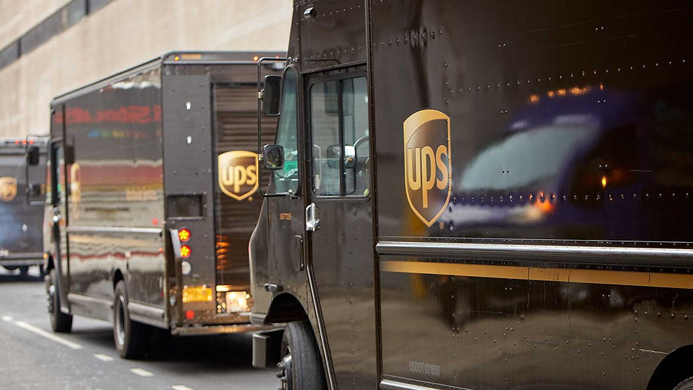 UPS trucks parked on a city street, displaying the company's brown and yellow logo. Buildings in the background, reflecting on truck surfaces.