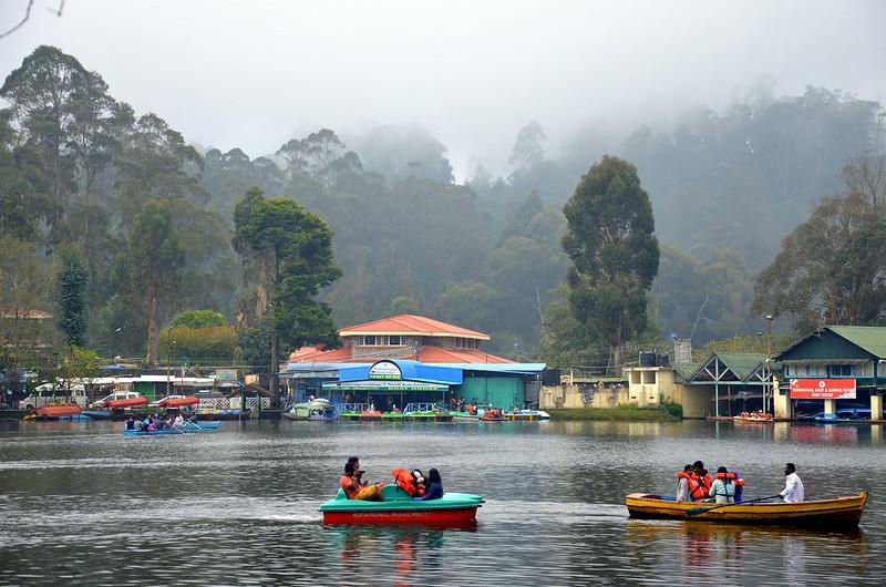 People in life jackets boating on a misty lake with colorful buildings and forested hills in the background.
