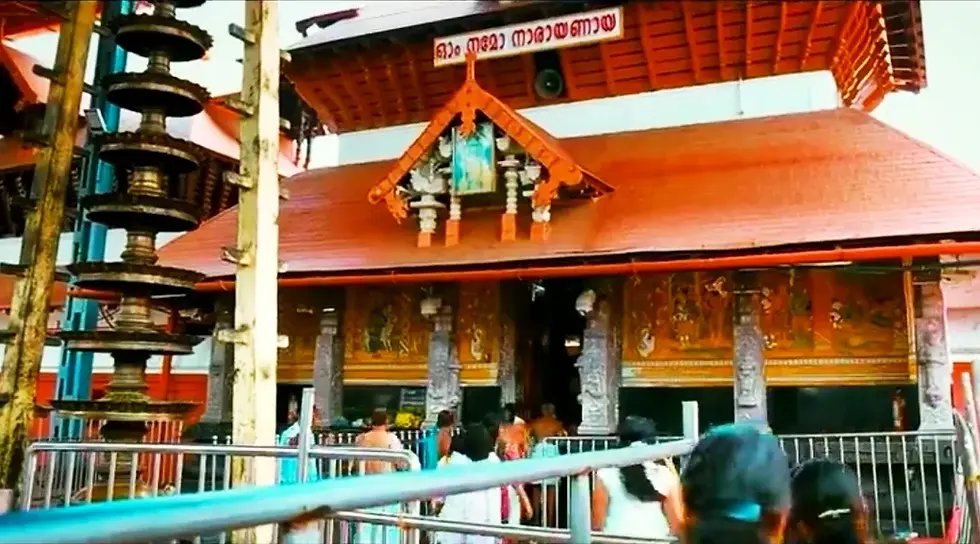 Crowd enters a red-roofed temple with decorative carvings. Brass lamps and a Malayalam sign are visible, creating a vibrant scene.