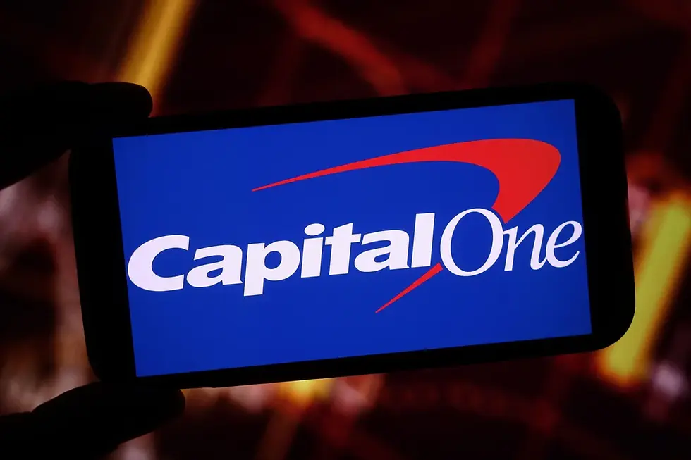 Smartphone displaying Capital One logo on blue screen, with a red abstract swoosh, against a blurred dark red and orange background.