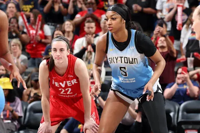 Two female basketball players face off; one in a red "Fever" jersey, the other in a blue "SkyTown" jersey. Crowd in background watches intently.