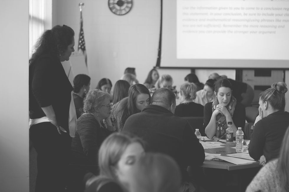 A group of people sit at tables in a classroom, engaged in discussion. A woman stands nearby. An American flag and a screen are visible.