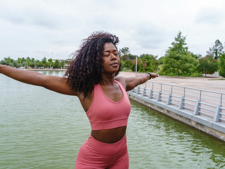 Black woman standing near a lake with arms extended and eyes closed.