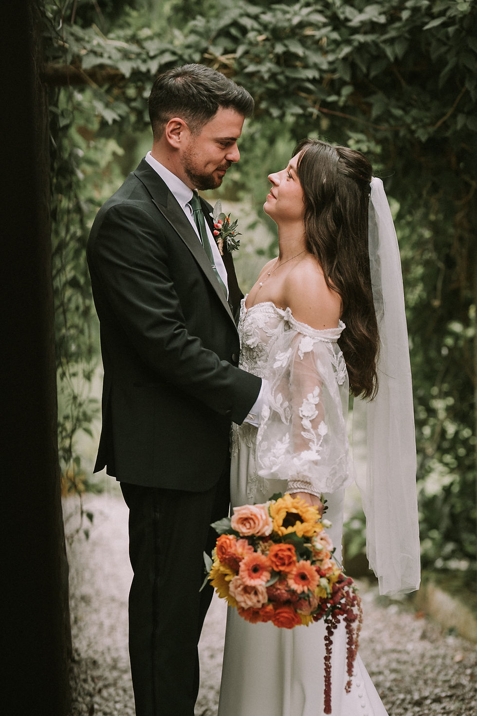 Bride and groom gaze at each other in a leafy garden, the bride holding a bright bouquet.