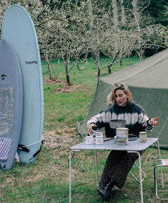En kvinna sitter och äter frukost vid ett bord utomhus, med blommande träd och surfbrädor i bakgrunden