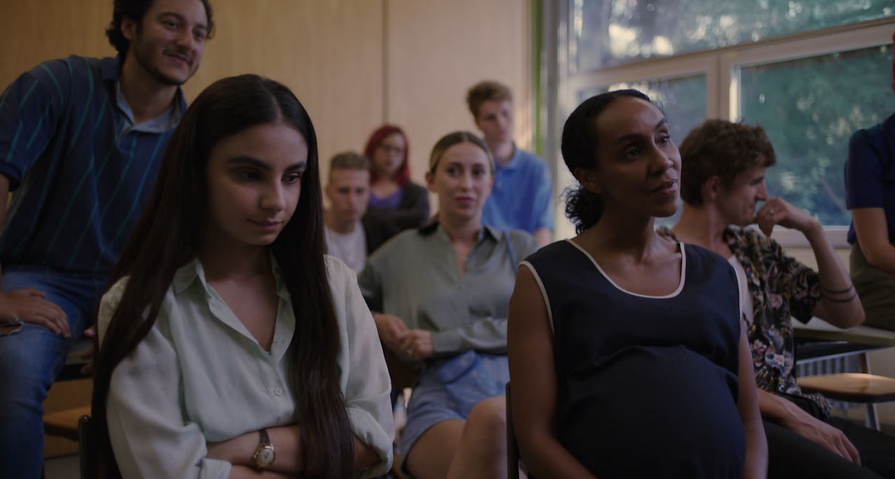 "Une jeune femme assise en classe, entourée d'étudiants, son expression sérieuse suggérant une scène académique du film 'ELAHA'
