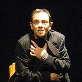 Un acteur passionné en costume rayé, Xavier Béja, incarne Jean Zay dans "L'Homme Complet", capturé en pleine performance, bras croisés et regard intense, évoquant un moment de profonde émotion.