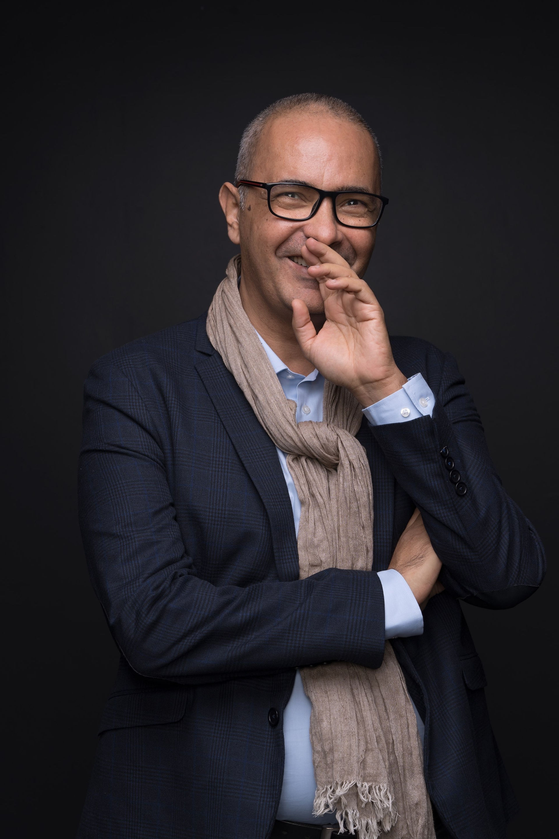 Portrait d'un homme aux lunettes, souriant et faisant un geste de la main près de sa bouche, donnant une impression de conversation informelle.