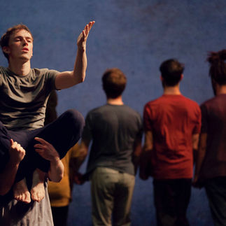 Un danseur soliste en équilibre précaire, jambes croisées en l'air, mains ouvertes et expressives, devant une ligne de danseurs flous en mouvement, symbolisant peut-être le leadership et la concentration au sein du collectif.