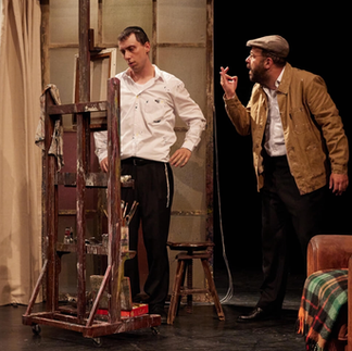 Deux acteurs sur scène dans 'Je m’appelle Asher Lev' : un homme en chemise blanche regarde un autre homme en costume et béret, qui tient une cigarette, à côté d'un chevalet de peinture, évoquant une discussion intense dans un atelier d'artiste.