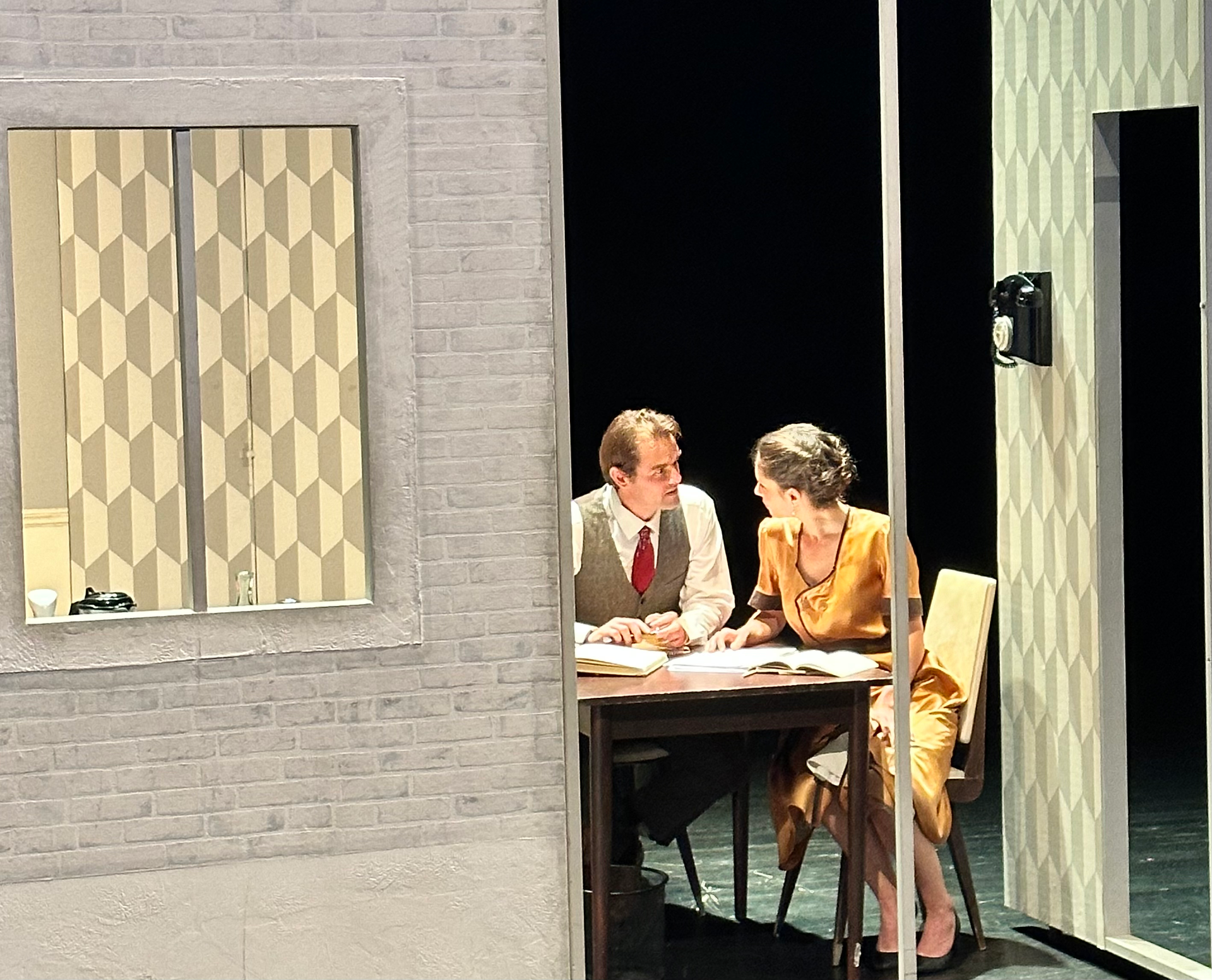 Deux acteurs plongés dans une conversation sérieuse sur scène, homme en costume avec cravate rouge et femme en robe orange satinée, dans un décor stylisé des années 50, évoquant une atmosphère intime et dramatique.