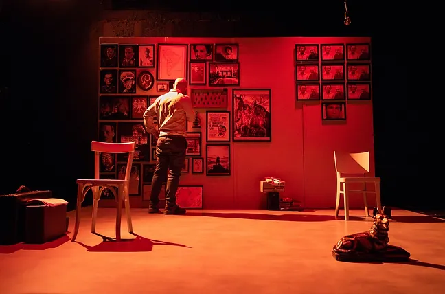 Scène du spectacle où Mikaël Chirinian se tient devant un mur couvert de photos et de documents, avec deux chaises blanches à ses côtés. L'éclairage rouge crée une atmosphère de tension et de confrontation.