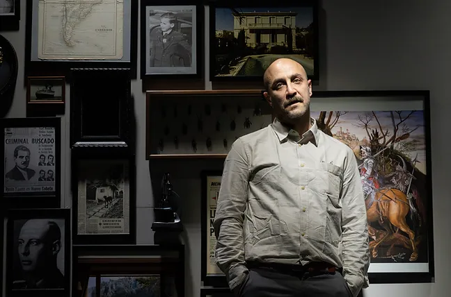Mikaël Chirinian debout, entouré de cadres et de documents relatifs à Josef Mengele. L'éclairage dramatique souligne l'atmosphère chargée de la pièce, accentuant le thème de la traque et de la mémoire historique.