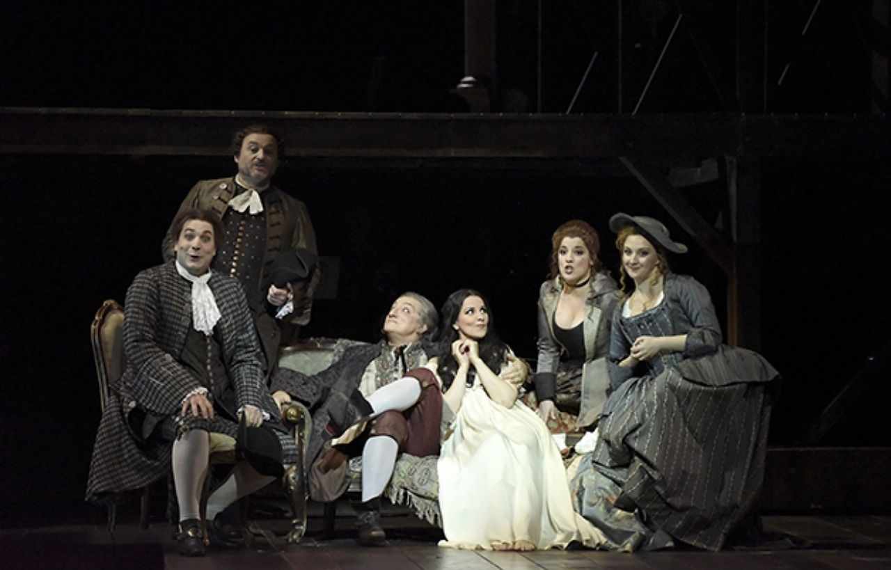 Acteurs en costumes du XVIIIe siècle rassemblés autour d'un canapé sur la scène de l'opéra 'Adriana Lecouvreur', évoquant une atmosphère intime et dramatique.