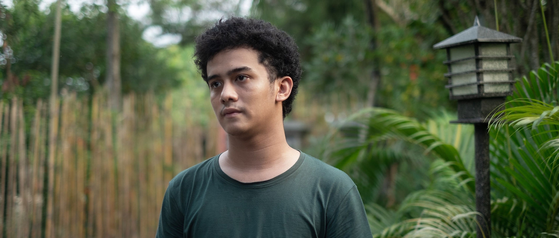 Un jeune homme en vert arbore un air pensif, debout à l'extérieur dans un cadre verdoyant, laissant présager une scène dans un environnement tropical luxuriant.