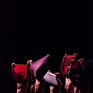 Danseurs en ligne, portant des vêtements décontractés dans des tons chauds, un danseur au centre s'étire vers l'arrière, dévoilant son torse nu sous une lumière teintée de bleu, évoquant un sentiment de libération et d'expression corporelle.
