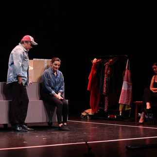 Sur scène, un acteur et une actrice en pleine action théâtrale, avec l'homme portant une casquette et la femme assise à côté d'une boîte marquée, suggérant une scène de la vie quotidienne ou une discussion importante.