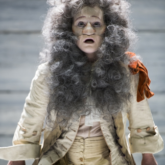 Acteur en costume baroque avec perruque grise bouclée et masque facial, incarnant un personnage extravagant de la pièce 'Le Mariage forcé' sur un fond de planches de bois