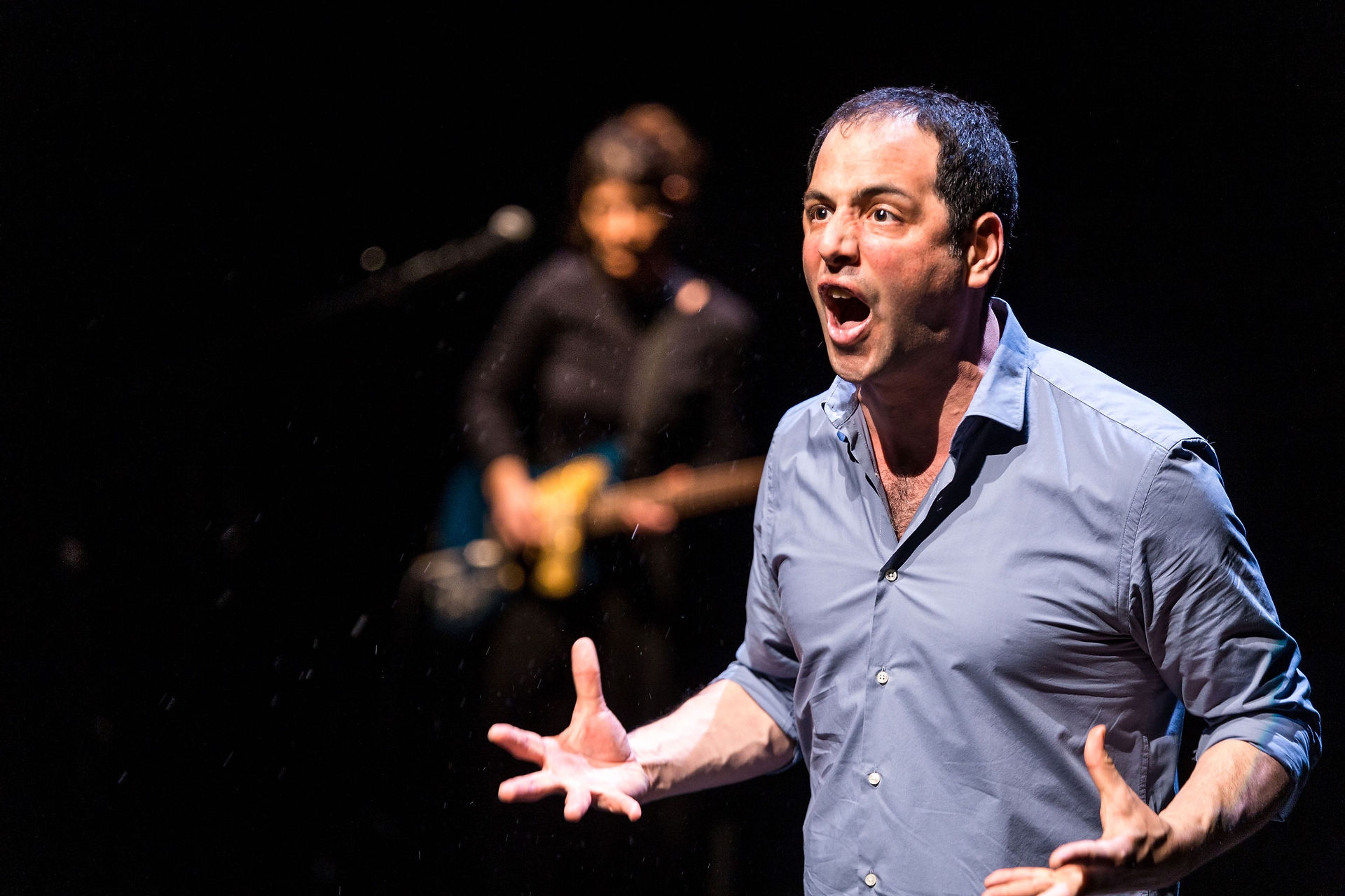Un acteur masculin sur scène, captivant et expressif, s'adresse passionnément au public avec un musicien flou en arrière-plan, illustrant l'union de la narration et de la musique en direct.